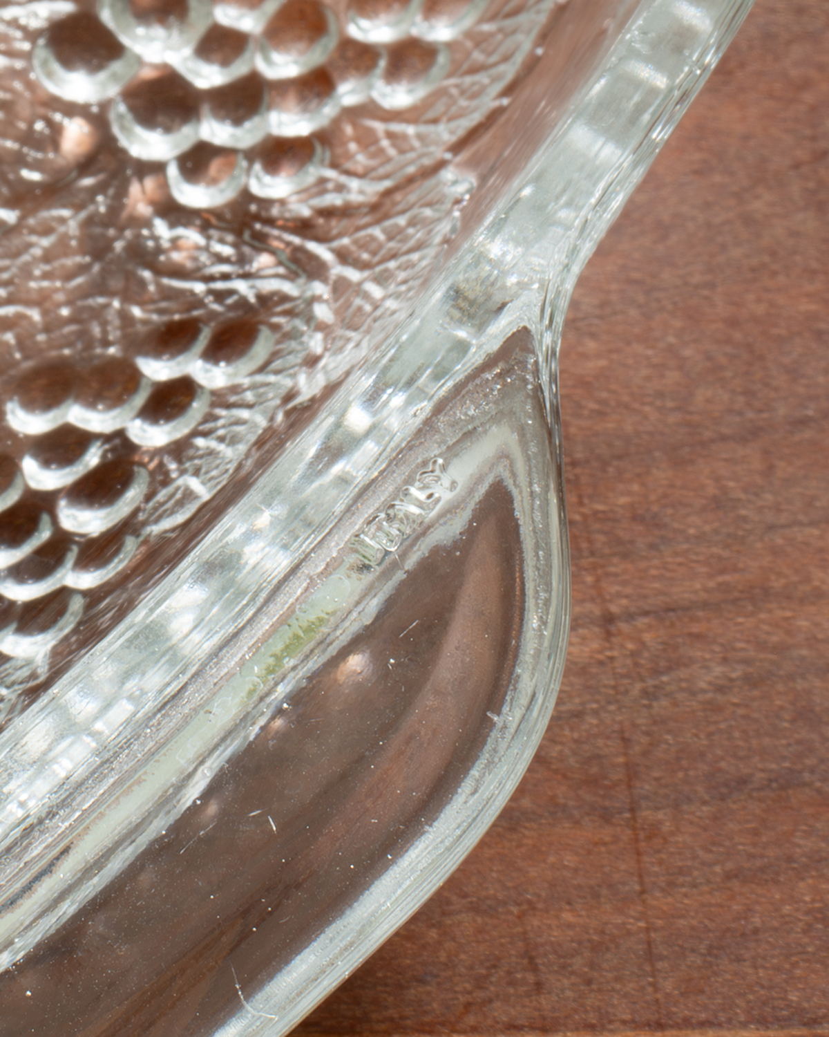 A close-up of a clear glass serving dish with a textured, embossed grape pattern on the top and a curved handle, resting on a brown wooden surface.