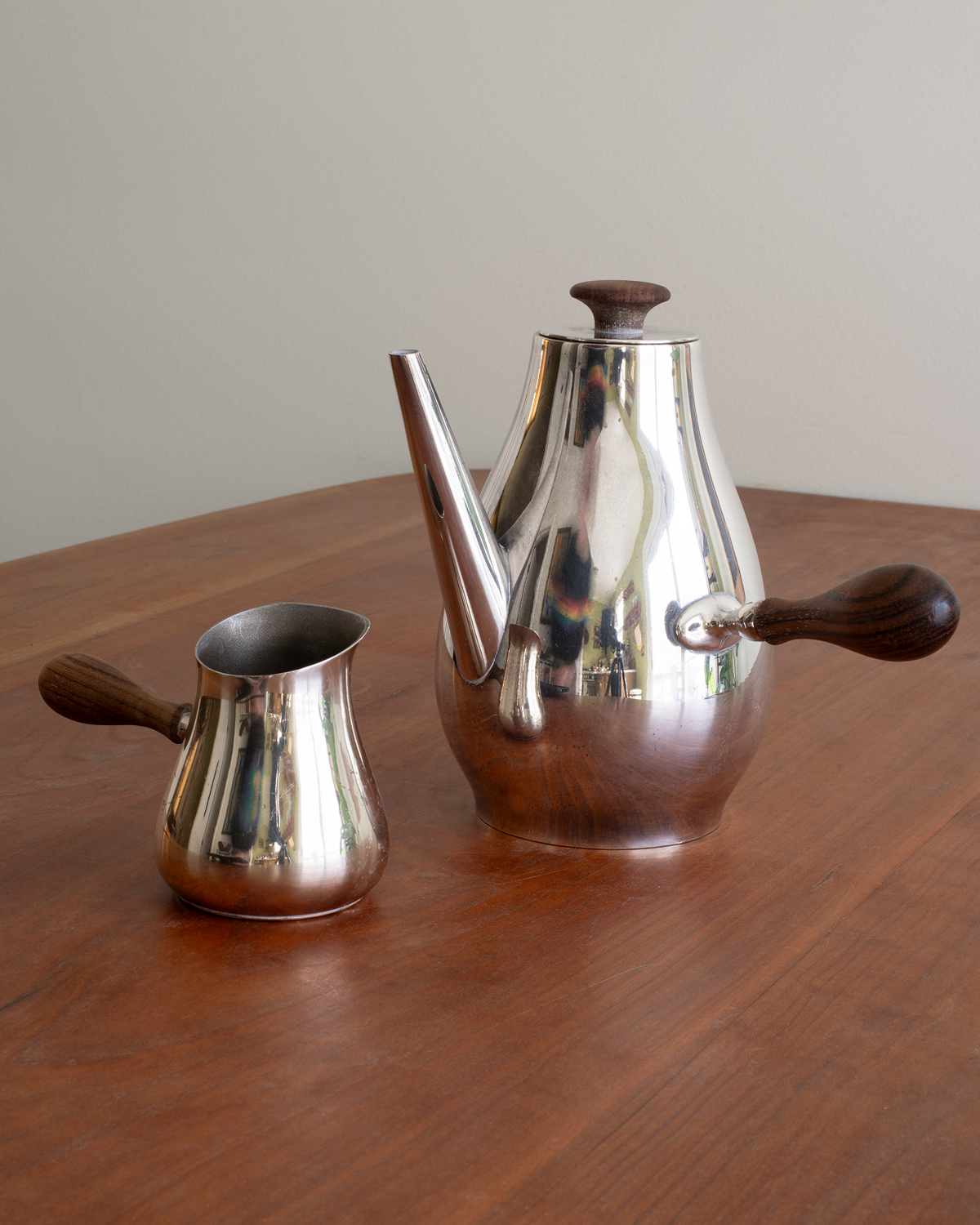 A polished silver metal coffee pot and matching milk pitcher with dark wooden handles, set on a wooden table against a plain light background.