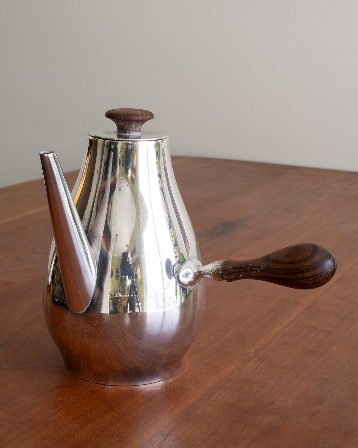 A polished silver metal coffee pot with dark brown wooden accents and handles, sitting on a wooden table against a neutral beige wall.