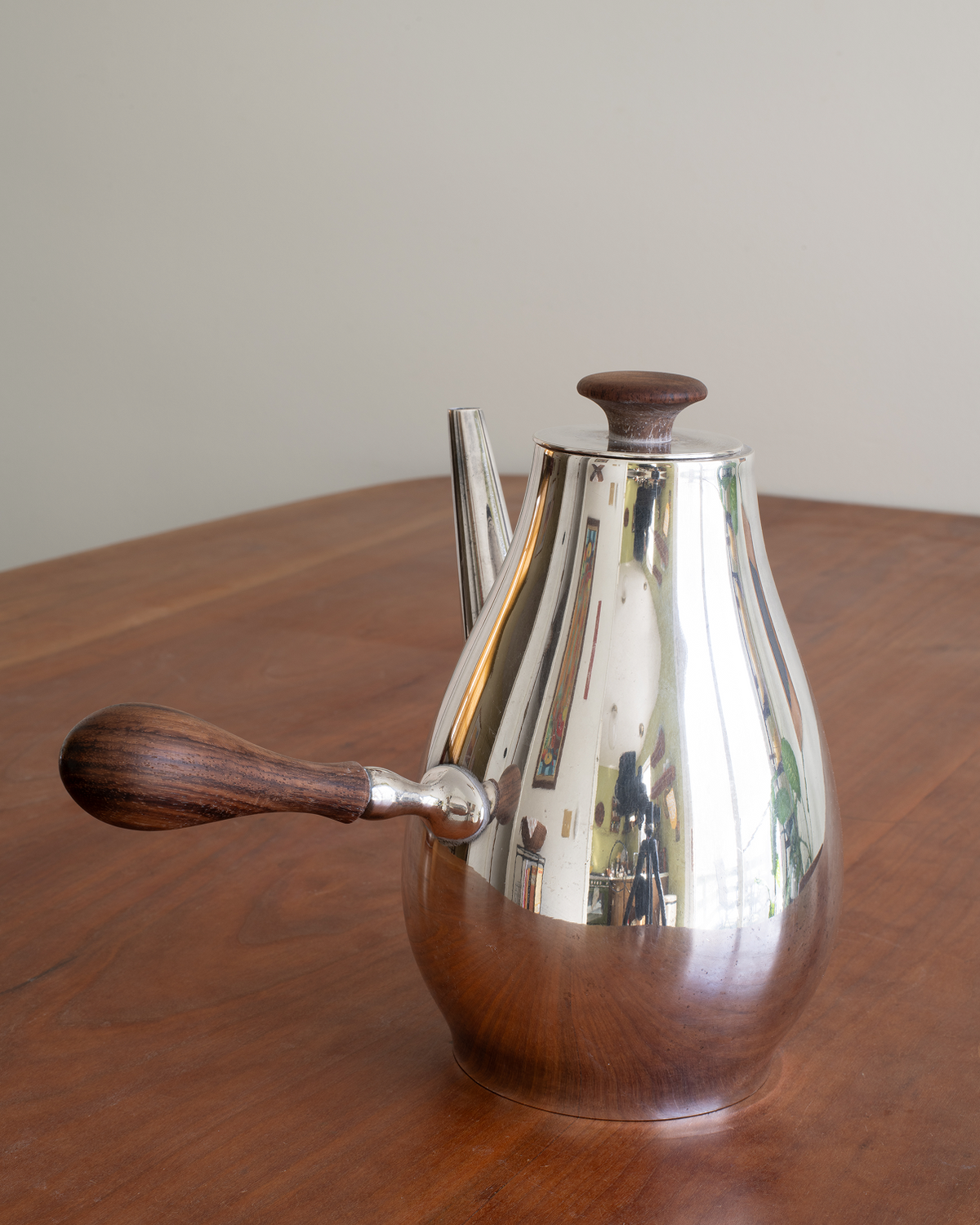 A polished silver metal coffee pot with dark wood handles and lid knobs, sitting on a wooden table against a plain beige wall.