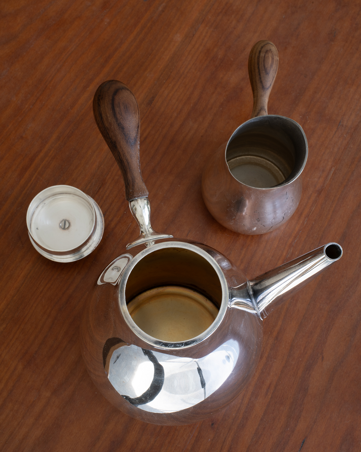 A vintage stainless steel teapot with a dark wood handle, shown with its lid off and a small matching metal cup on a wooden tabletop.