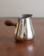 A polished silver metal coffee pitcher with a dark wooden handle, sitting on a wooden table against a neutral beige background.