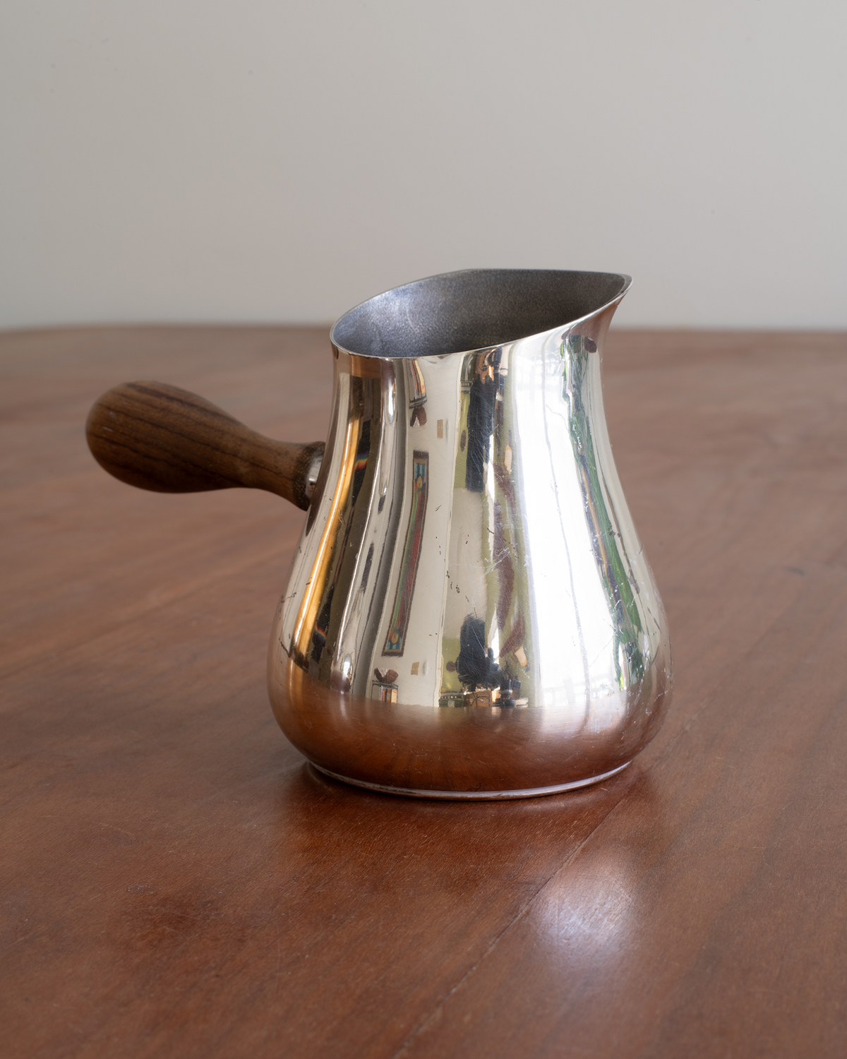 A polished silver metal coffee pitcher with a dark wooden handle, sitting on a wooden table against a neutral beige background.