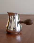 A polished metal Turkish coffee pot with a dark wood handle, sitting on a wooden table against a plain light background.