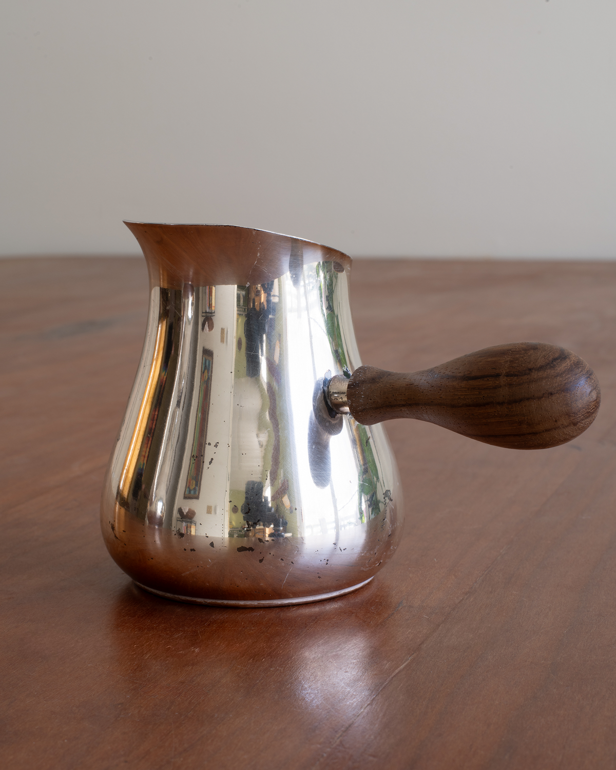 A polished metal Turkish coffee pot with a dark wood handle, sitting on a wooden table against a plain light background.
