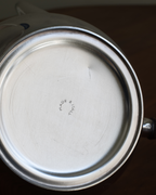Close-up bottom view of a brushed stainless steel pot or kettle base with a small stamped 'MADE IN ITALY' mark in the center, set against a dark brown background.