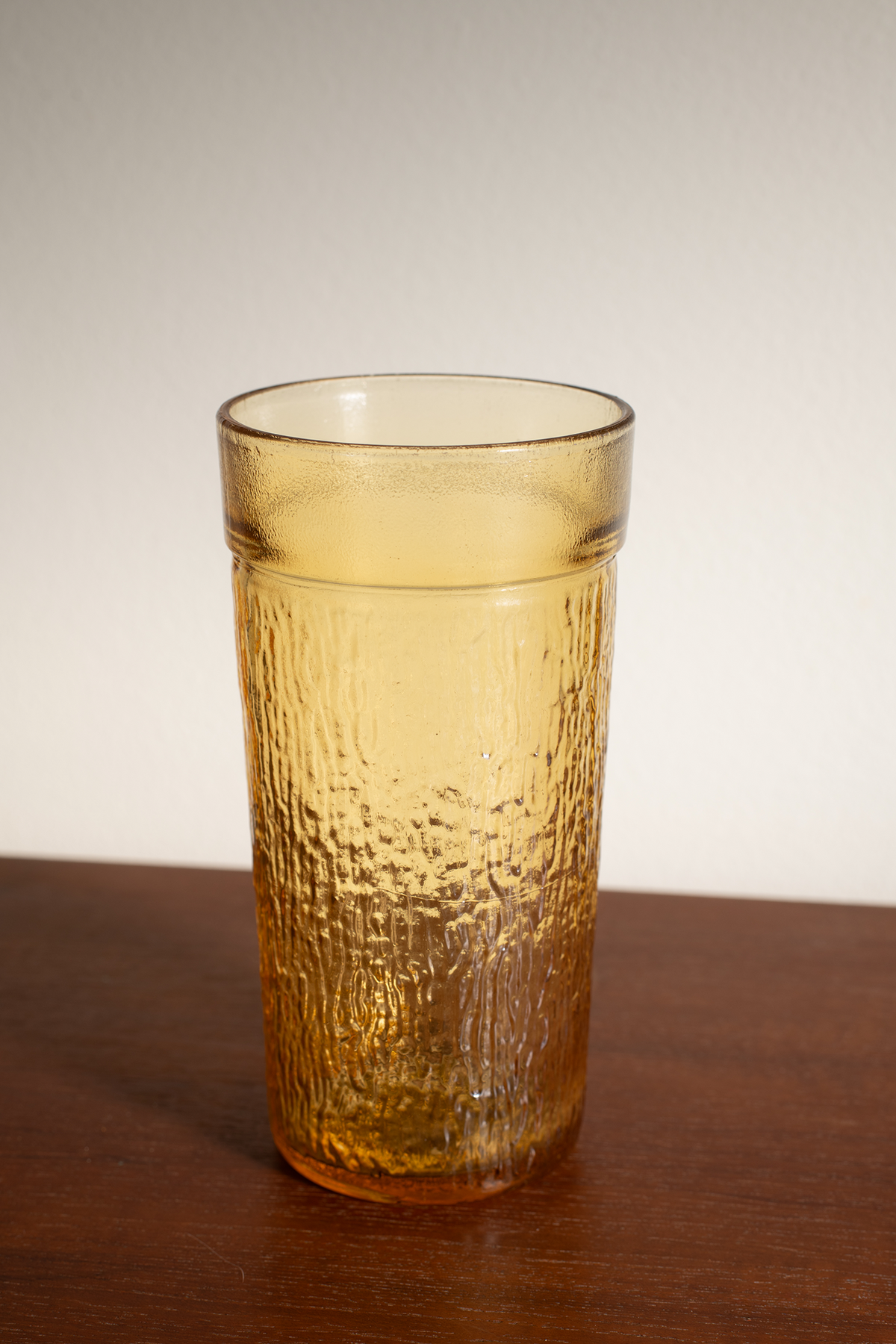 A textured amber glass tumbler with a hammered finish, standing on a dark wooden surface against a neutral light gray background.
