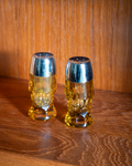 Two small skull-shaped beer glasses with metallic silver caps, filled with amber beer, set on a wooden table against a wooden wall.