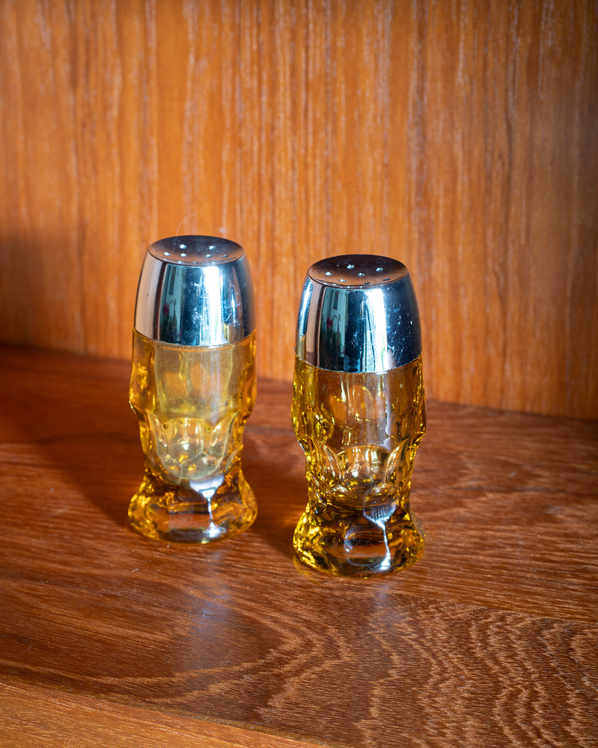 Two small skull-shaped beer glasses with metallic silver caps, filled with amber beer, set on a wooden table against a wooden wall.
