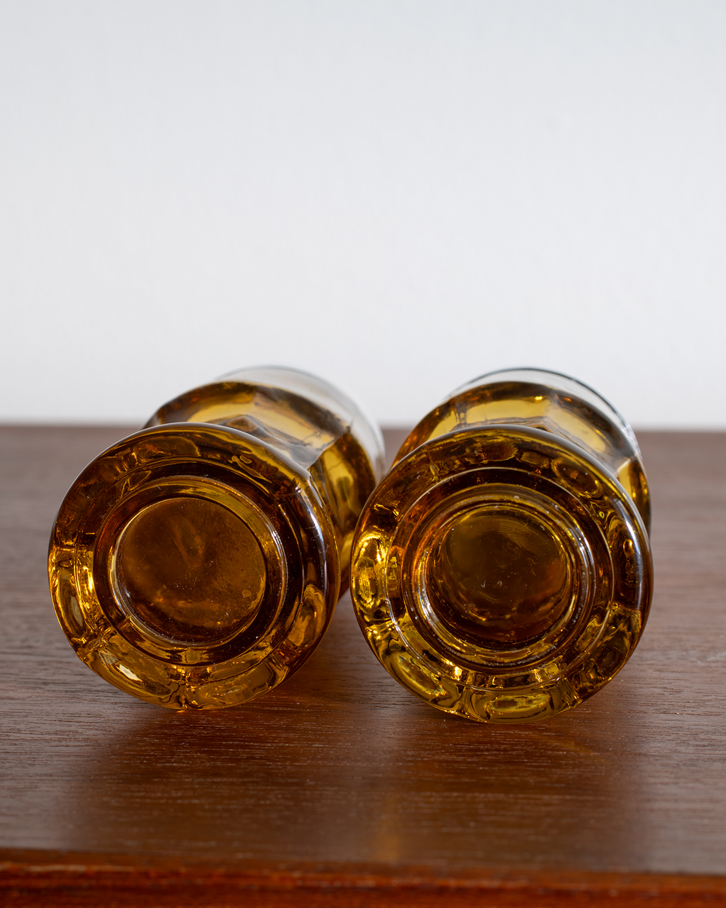 Two amber glass bottle bottoms or decorative glass pieces lying side by side on a dark wooden surface against a plain light background.