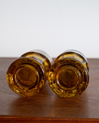Two amber glass bottle bottoms or decorative glass pieces lying side by side on a dark wooden surface against a plain light background.