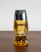 A skull-shaped amber glass tumbler with a polished silver metallic lid, set on a wooden tabletop against a neutral light gray background.