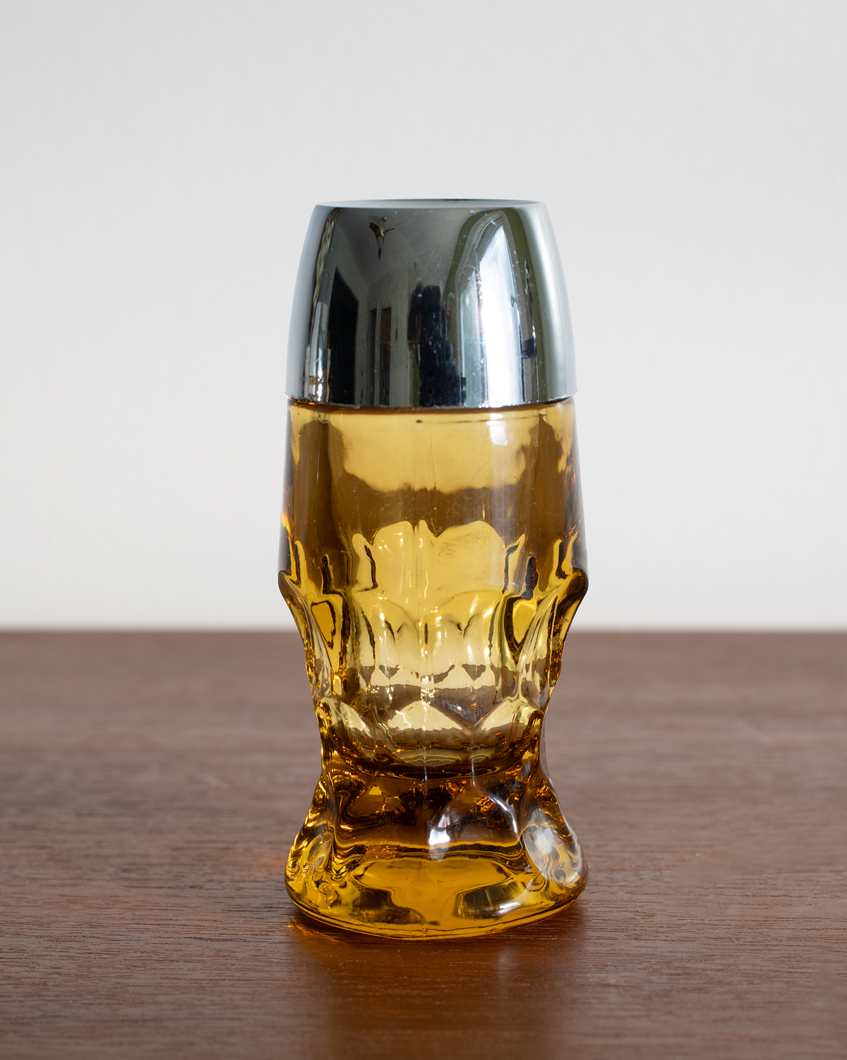 A skull-shaped amber glass tumbler with a polished silver metallic lid, set on a wooden tabletop against a neutral light gray background.