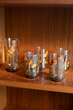 Five clear glass tumblers with etched leaf patterns and orange butterfly designs, displayed on a wooden shelf.