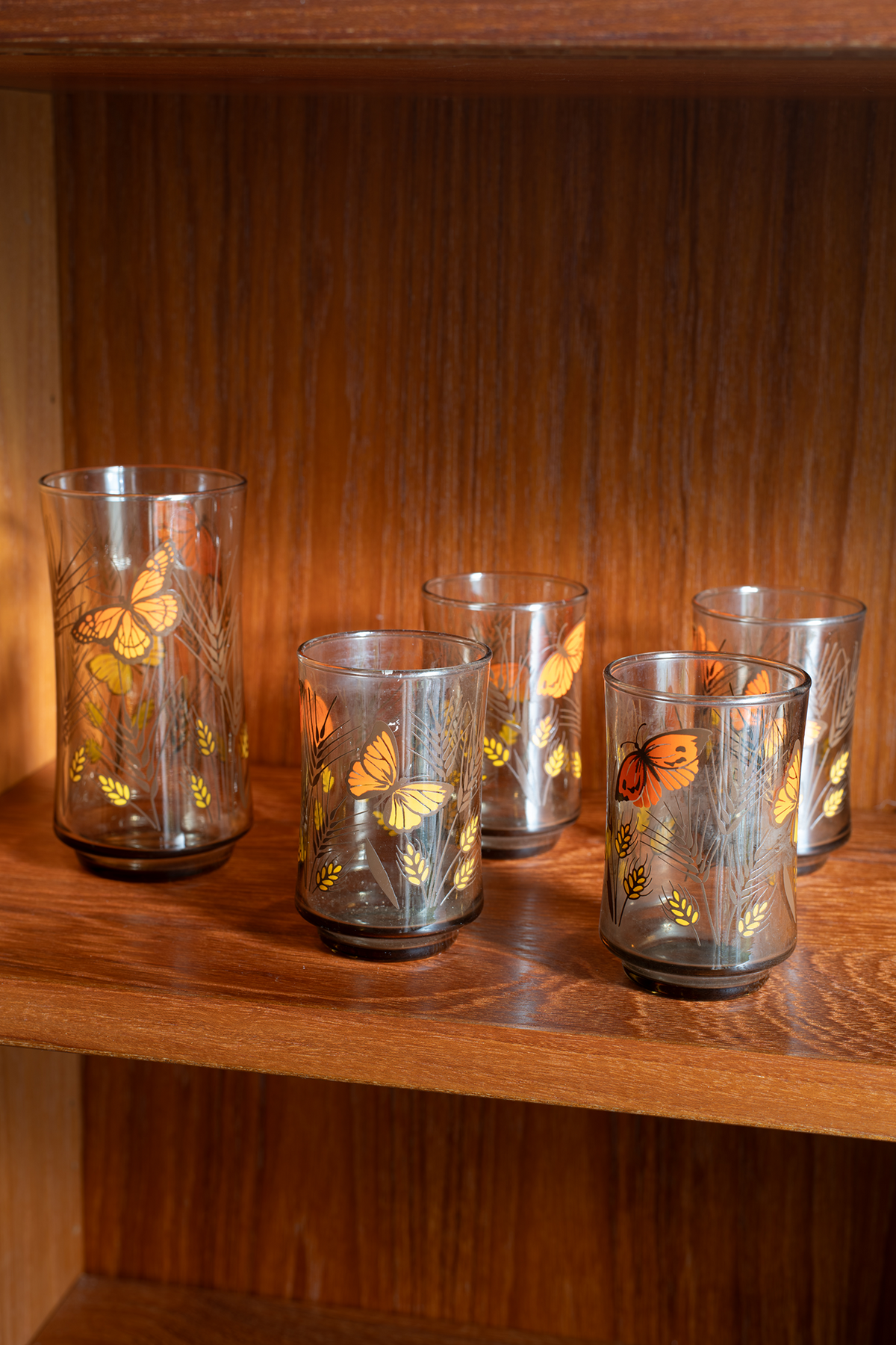 Five clear glass tumblers with etched leaf patterns and orange butterfly designs, displayed on a wooden shelf.