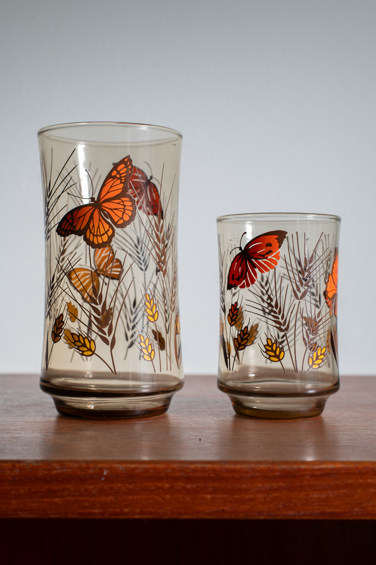 Two clear glass tumblers with vintage-style orange and yellow butterfly and leaf illustrations, displayed on a wooden surface against a neutral gray background.