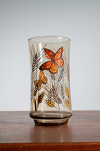 A vintage-style clear glass tumbler with a brown tint, decorated with orange butterflies and yellow wheat stalks, resting on a wooden surface against a light gray background.