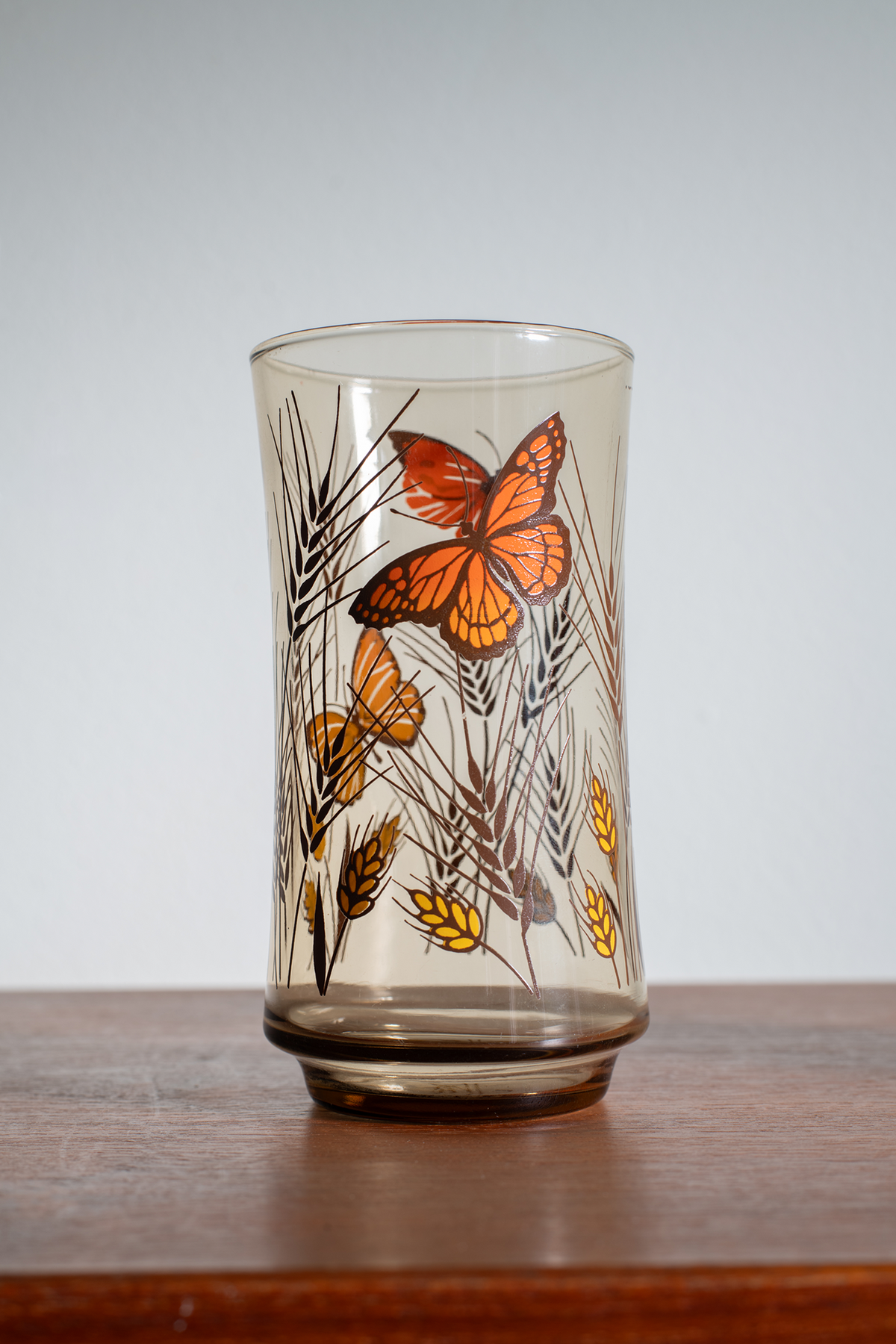 A vintage-style clear glass tumbler with a brown tint, decorated with orange butterflies and yellow wheat stalks, resting on a wooden surface against a light gray background.