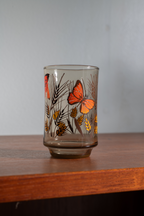 A clear glass tumbler with a retro design featuring orange butterflies and gold wheat stalks on a brown background, sitting on a wooden shelf against a gray wall.