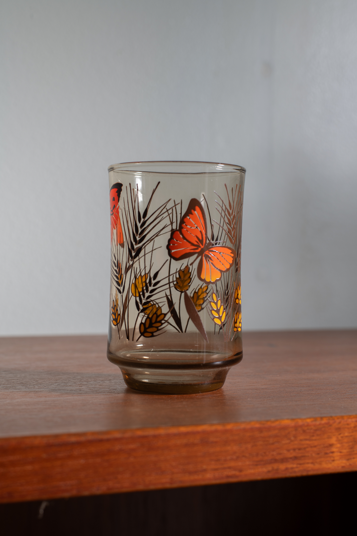 A clear glass tumbler with a retro design featuring orange butterflies and gold wheat stalks on a brown background, sitting on a wooden shelf against a gray wall.
