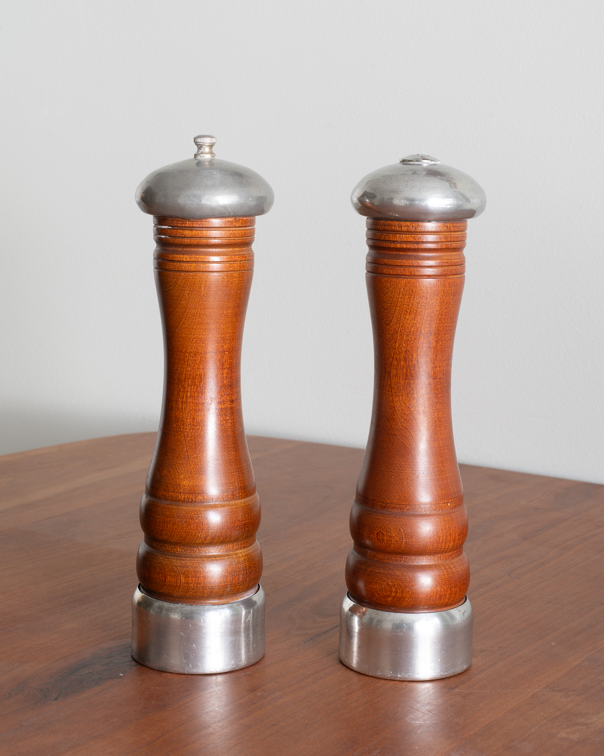 Two wooden pepper and salt mills with polished silver metal caps and bases, standing on a wooden table against a light gray background.