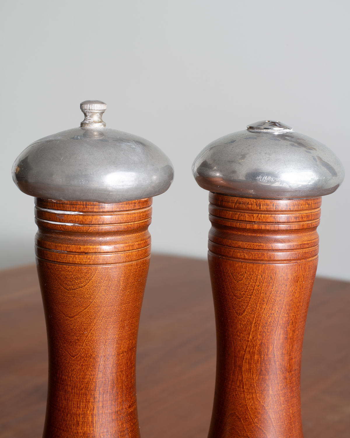 Two wooden pepper and salt mills with polished silver metal tops, shown on a wooden surface against a neutral gray background.