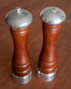 A pair of vintage wooden pepper and salt mills with polished silver metal tops and bases, set against a warm brown wooden background.