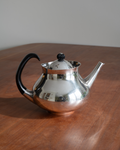 A polished silver teapot with a black curved handle and lid knob, sitting on a wooden table against a neutral gray background.