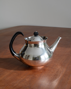 A polished silver teapot with a black curved handle and lid knob, sitting on a wooden table against a neutral gray background.