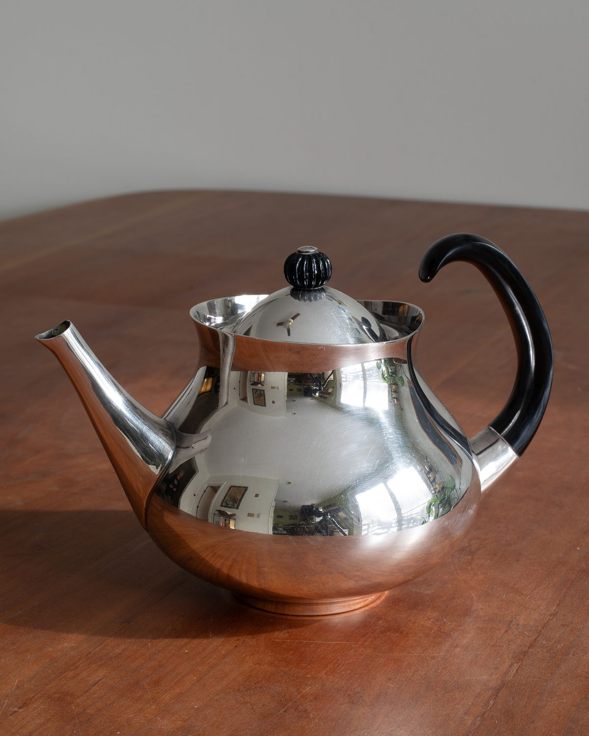 A polished silver teapot with a black handle and lid knob, sitting on a wooden table with a neutral gray background.