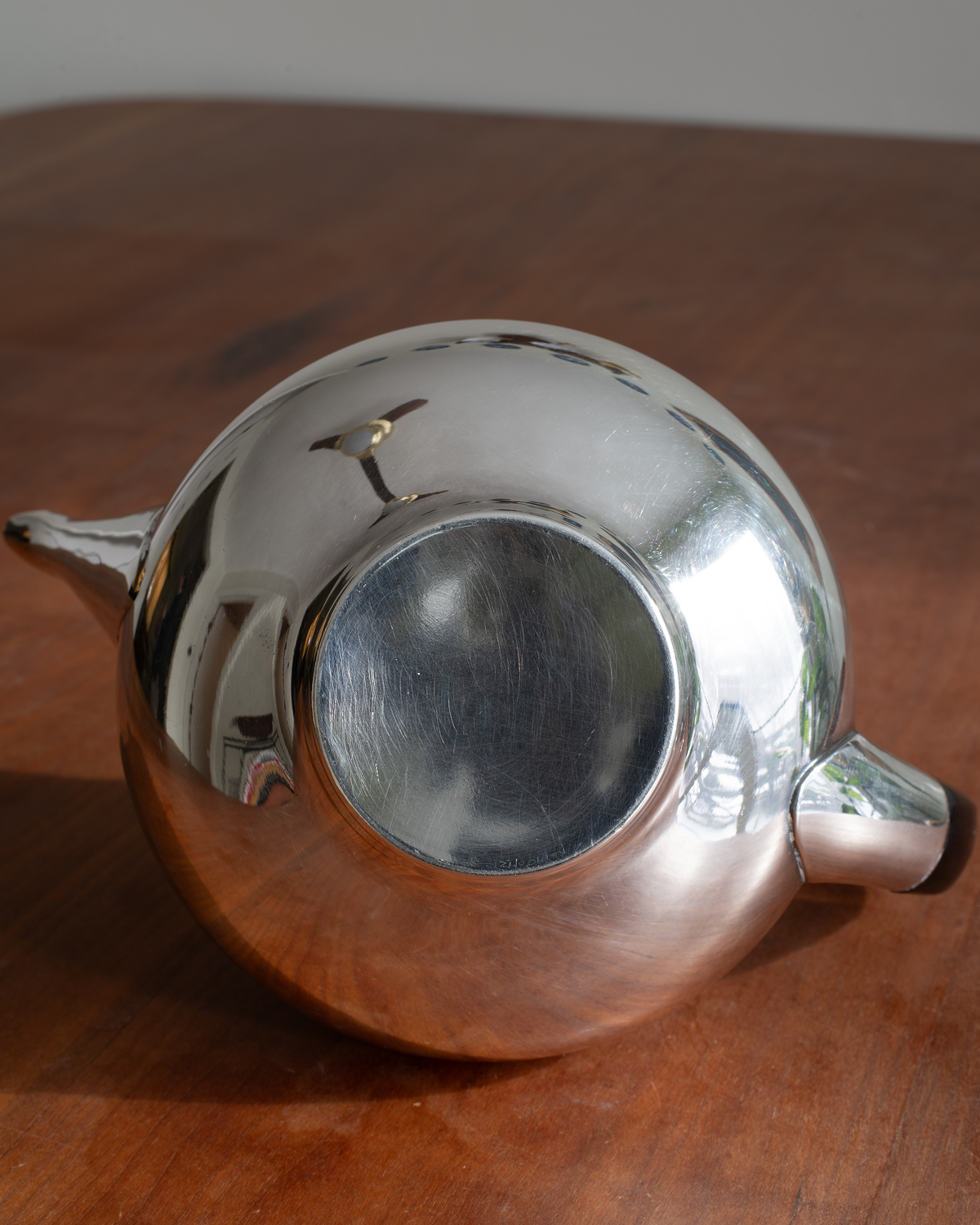 A polished silver teapot with a highly reflective metallic surface, shown from a top-down angle on a wooden tabletop.