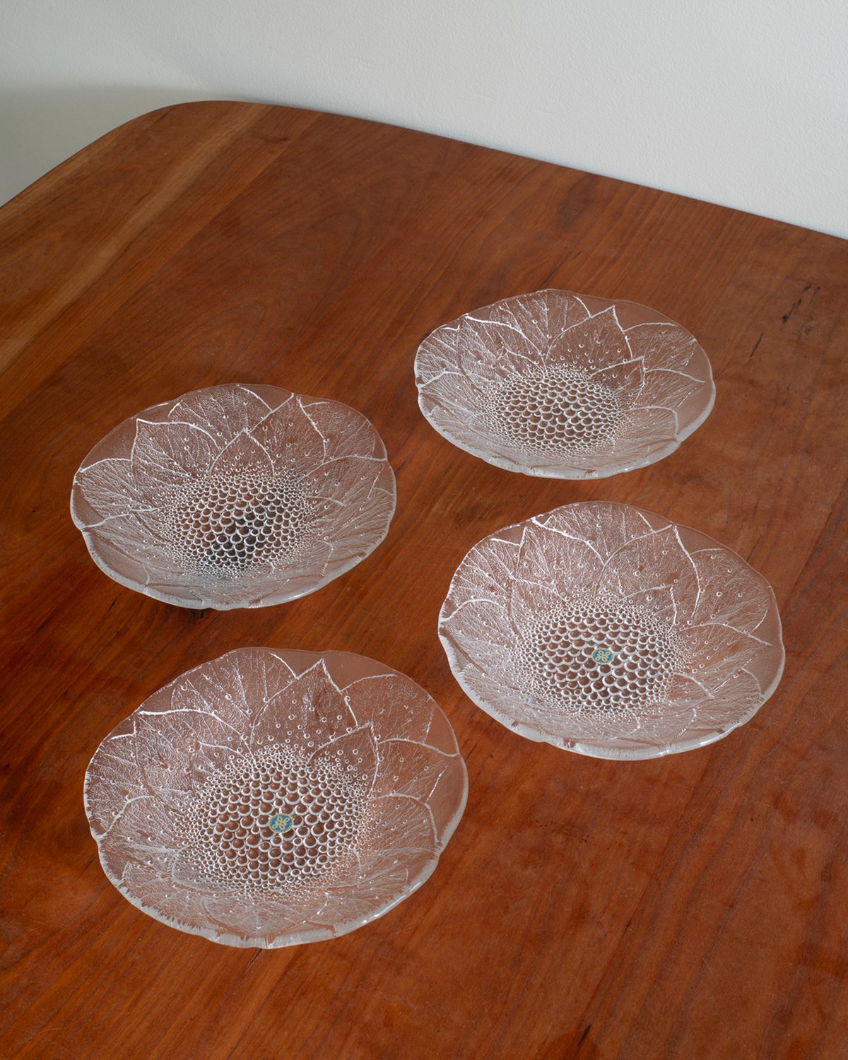 Four clear textured glass plates arranged on a warm brown wooden tabletop against a light neutral wall.