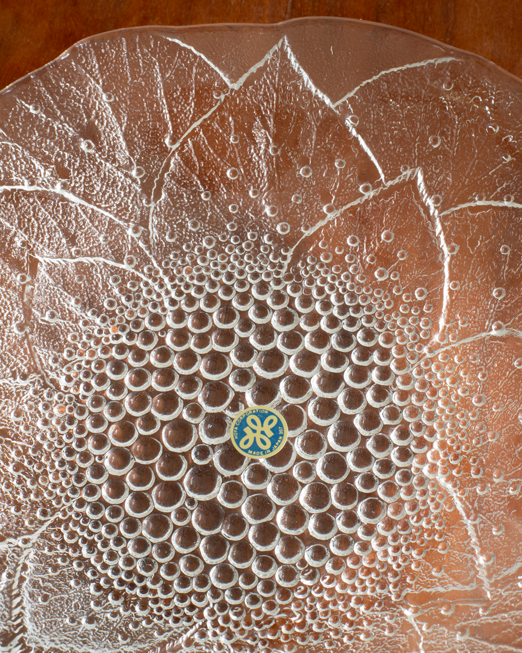 A close-up of a clear textured glass plate with a raised floral petal pattern and a small blue and gold circular logo at the center, photographed on a brown wooden surface.