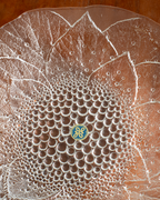 A close-up of a clear textured glass plate with a raised floral petal pattern and a small blue and gold circular logo at the center, photographed on a brown wooden surface.