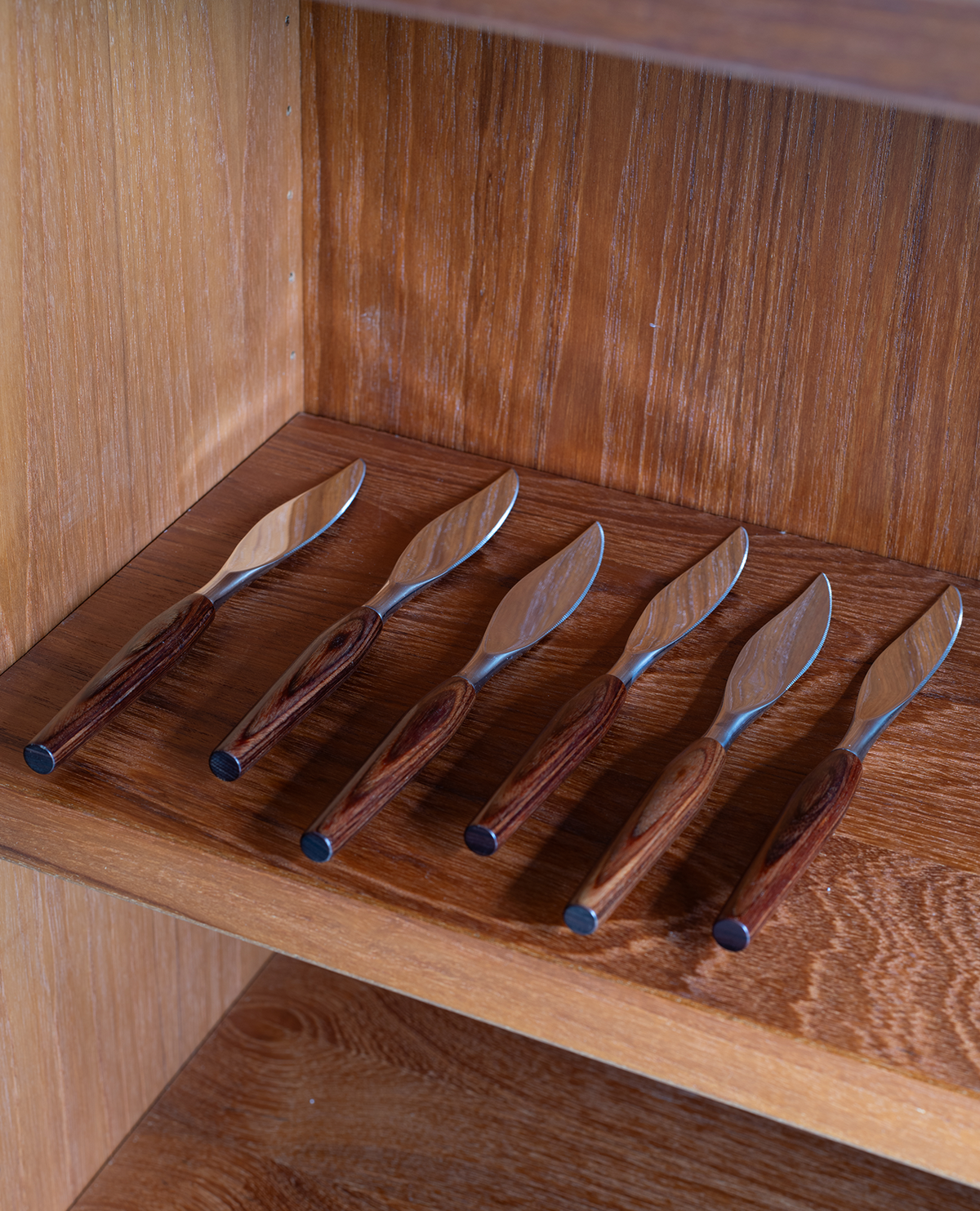 Five wooden spreader knives with dark wood handles and silver metal blades, arranged diagonally on a wooden shelf inside a wooden cabinet.