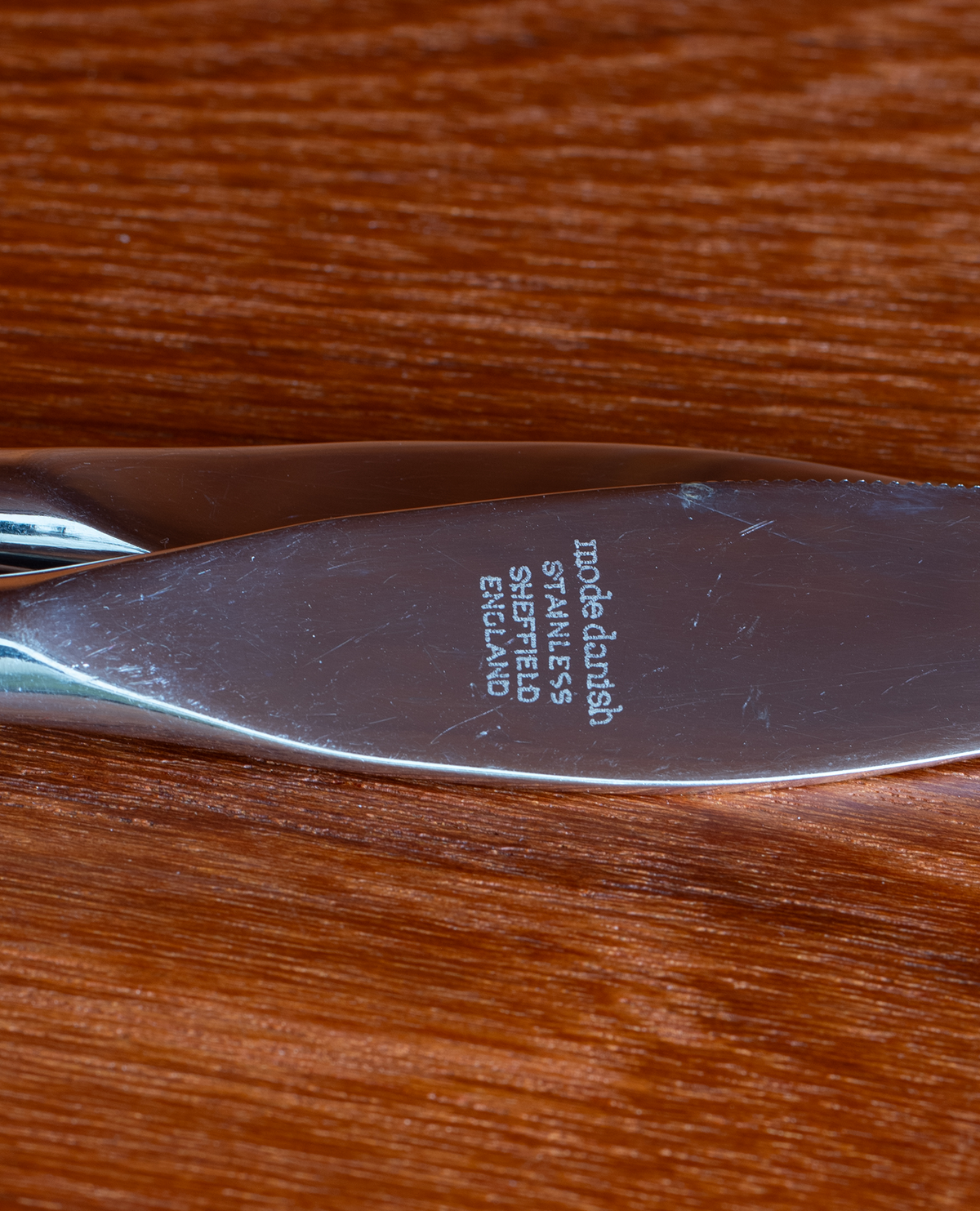 Close-up of a stainless steel knife blade resting on a warm brown wooden surface, with engraved text reading 'modelladish STAINLESS STEEL SHIPFIELD EDITION'.