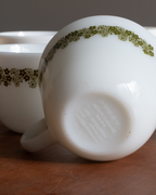 A close-up of a white milk glass mug with a green floral pattern band, shown upside down on a wooden surface with another mug blurred in the background.