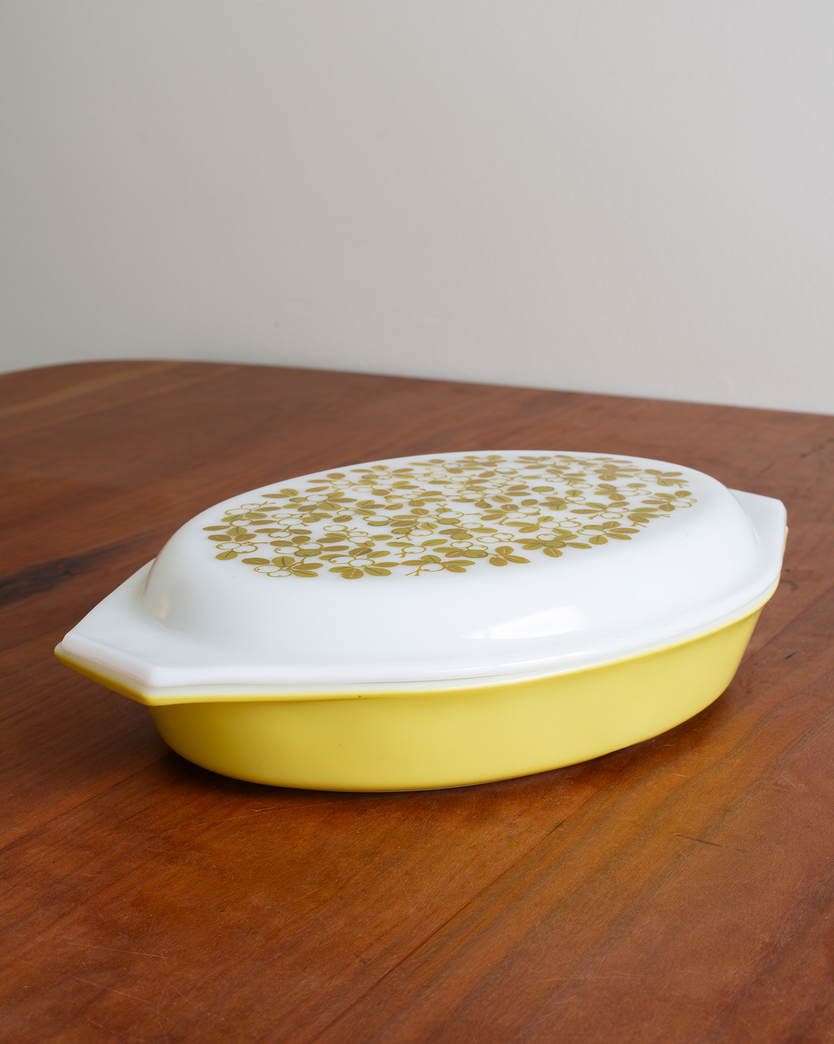 A yellow oval casserole dish with a white lid decorated with a gold floral pattern, resting on a wooden table against a plain wall.