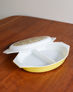 A yellow and white divided ceramic dish with a matching white lid featuring a gold speckled pattern, resting on a wooden table.