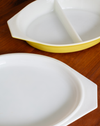 A white ceramic divided serving dish with a yellow rim, shown on a wooden surface with its matching white lid in the foreground.