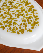 A close-up of a white oval plate or dish featuring a repeating olive-green floral pattern, resting on a brown wooden surface.