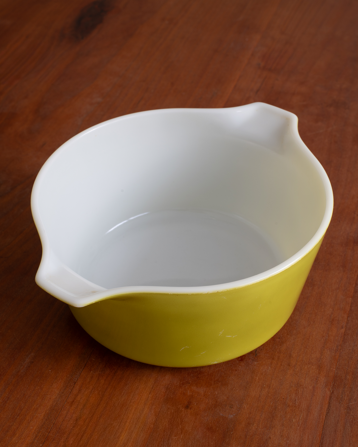 A small ceramic mixing bowl with a yellow exterior and white interior, featuring integrated pouring spouts, shown on a wooden surface.