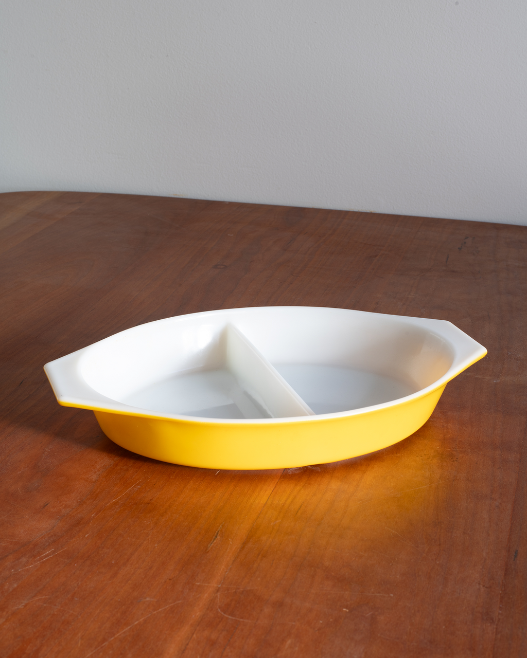A yellow and white ceramic divided serving dish with side handles, placed on a wooden table against a plain wall.