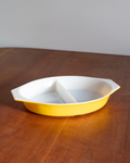 A yellow and white ceramic divided serving dish with side handles, placed on a wooden table against a plain wall.