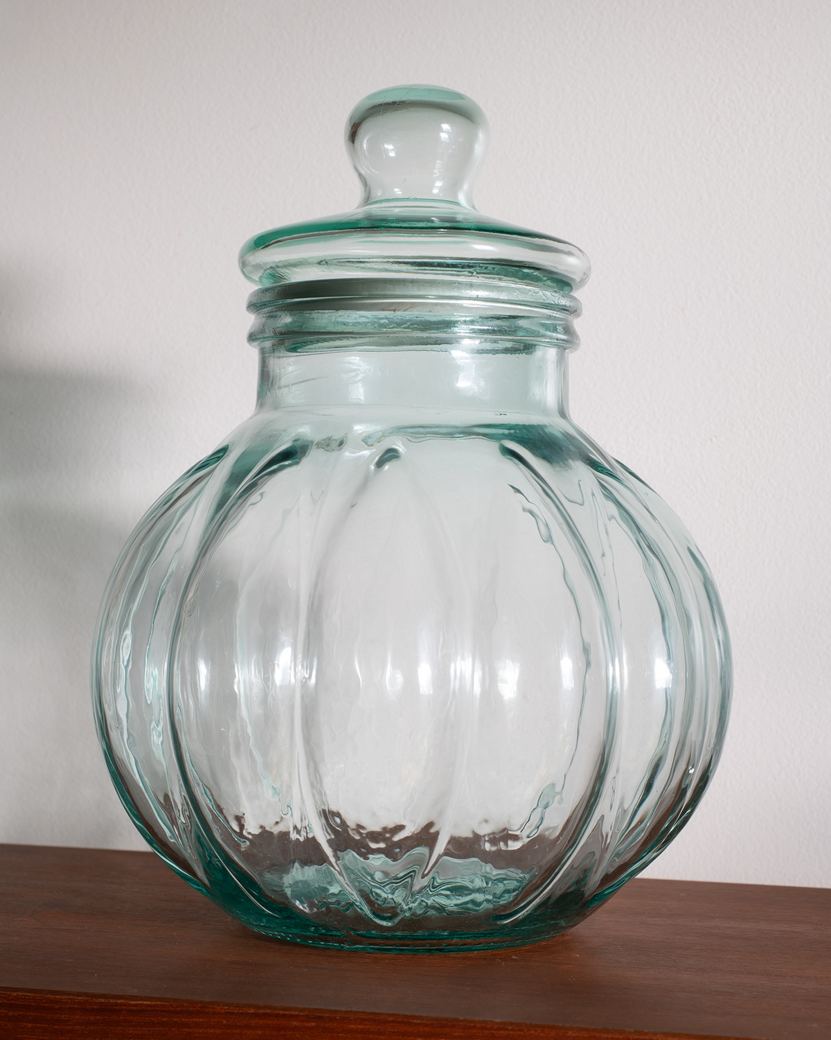 A clear glass jar with a matching glass lid, featuring a ribbed, pumpkin-like shape and a small knob on top, displayed on a wooden surface against a plain white wall.