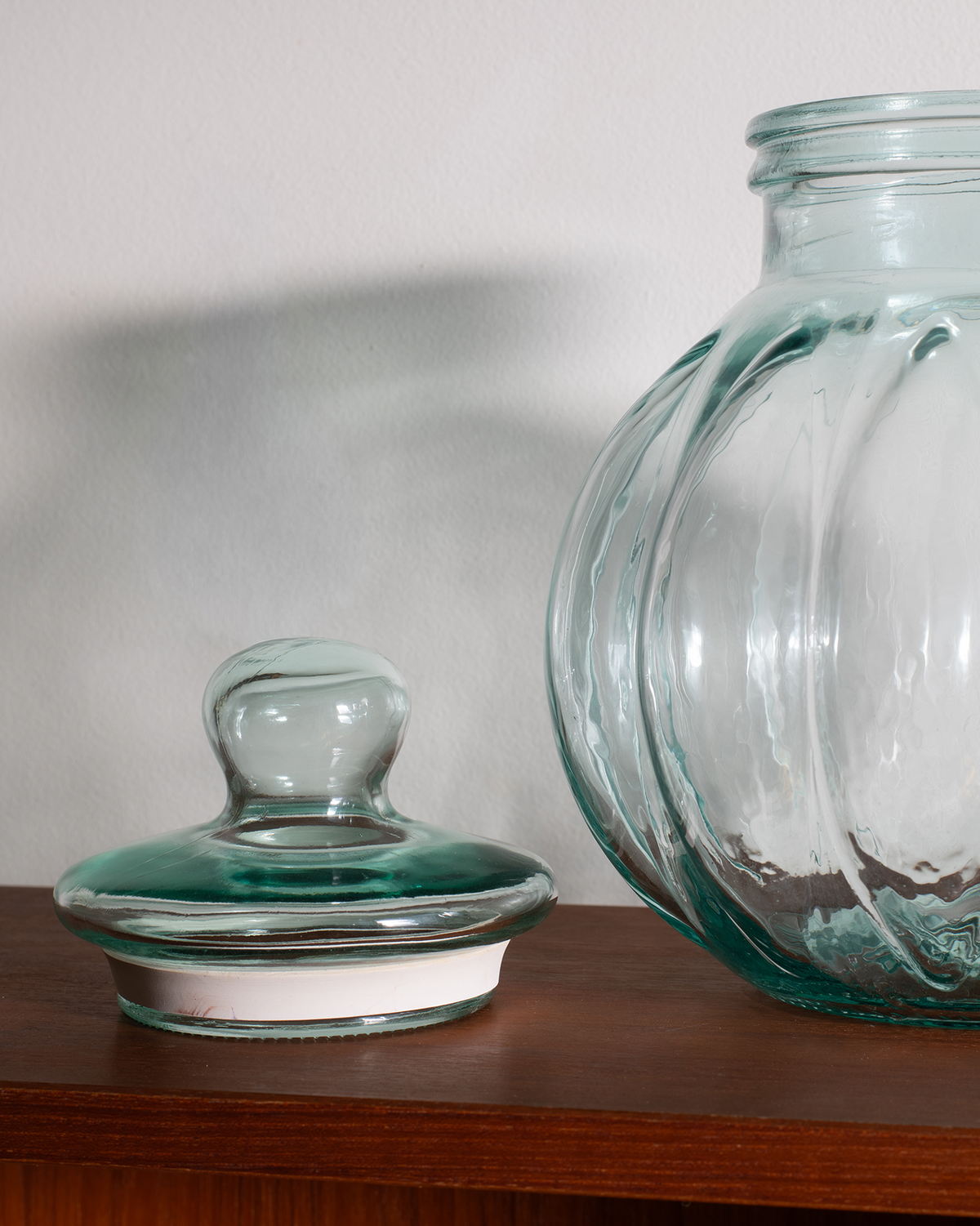 A close-up of a clear glass jar with a matching glass lid resting on a wooden shelf against a plain light wall.