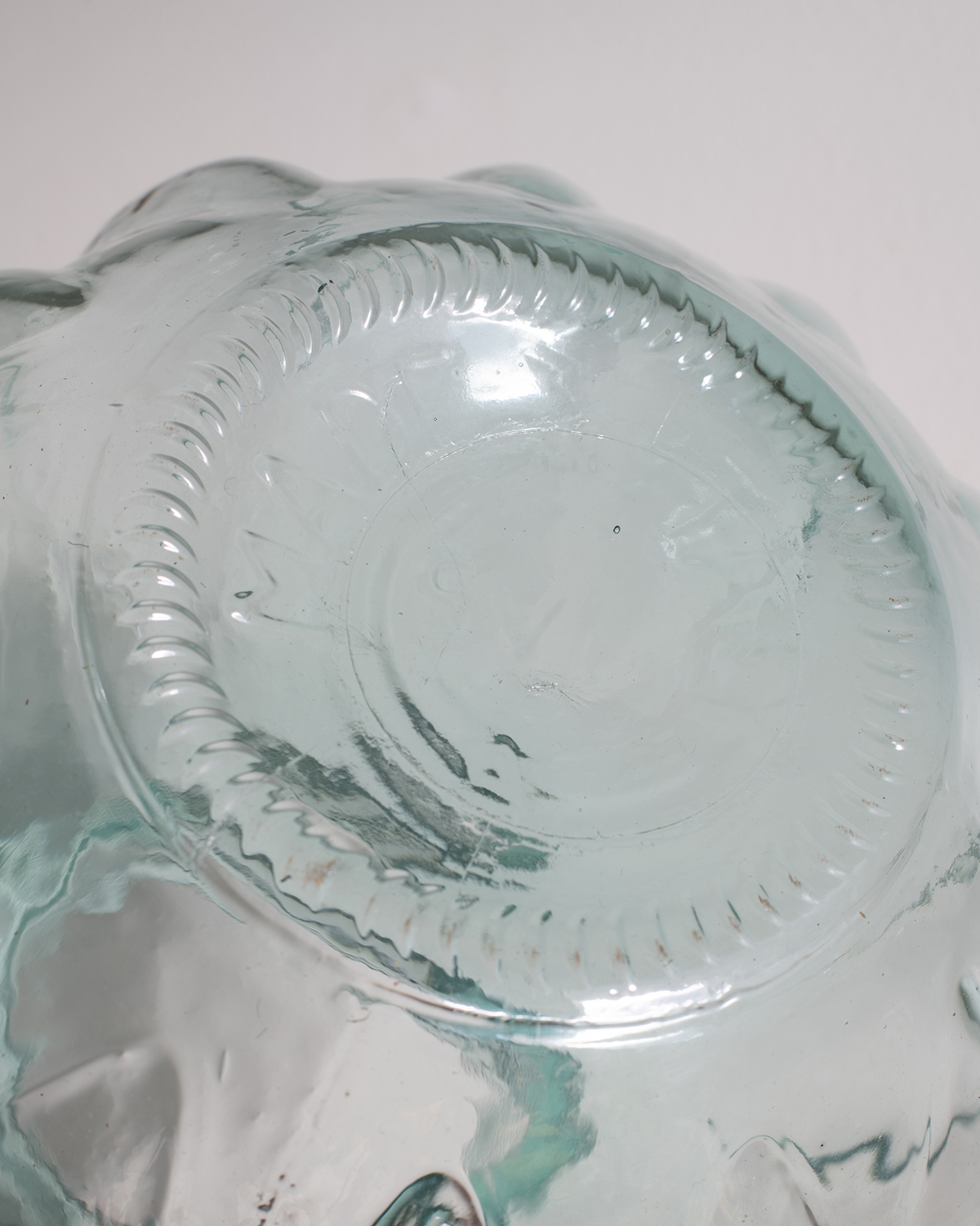 A close-up of the bottom of a clear glass bowl or pitcher with a fluted rim and embossed circular detail, showing a light blue-green tint in the glass against a white background.