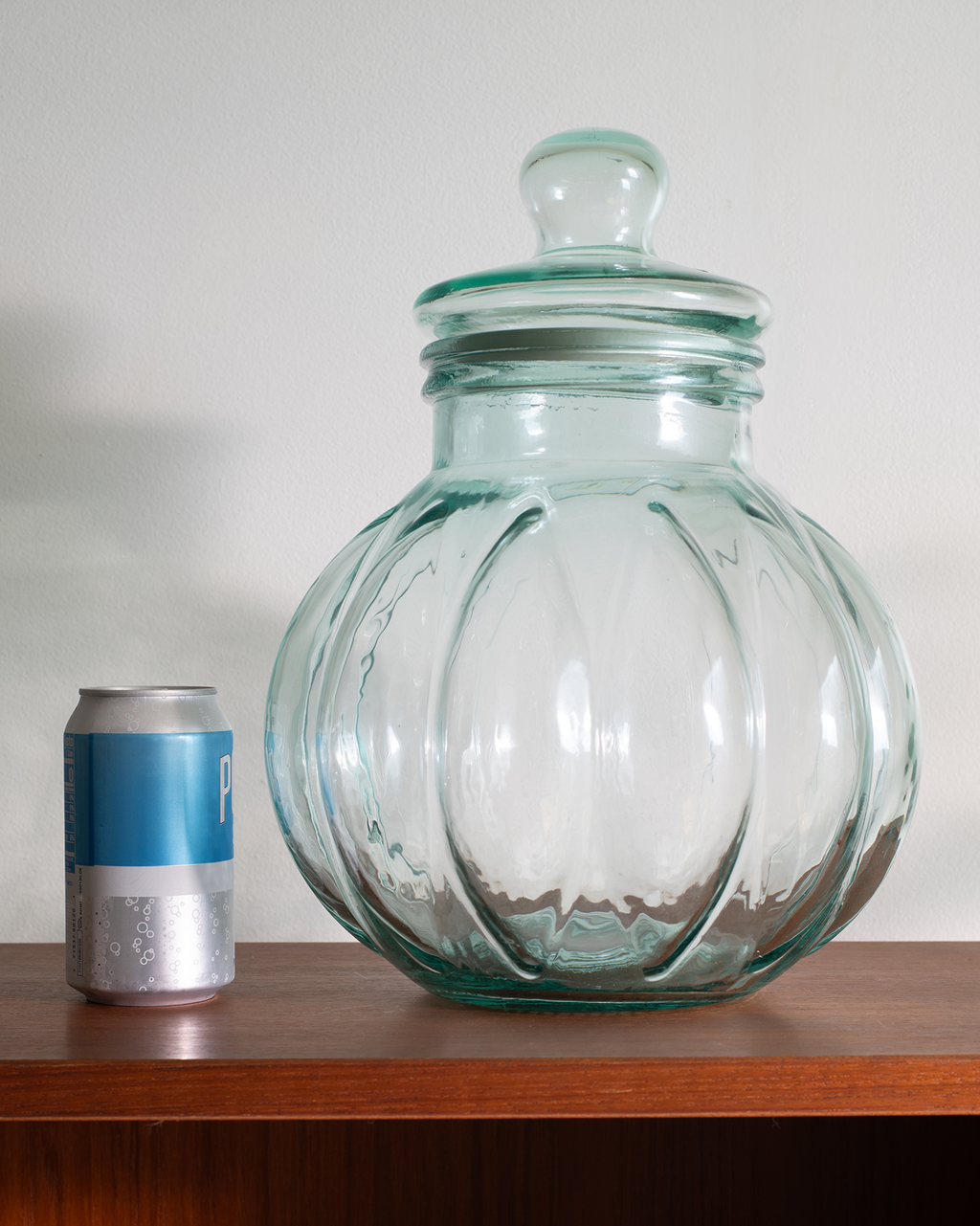 A large clear glass jar with a matching lid, featuring a ribbed, pumpkin-like design, displayed on a wooden shelf next to a soda can for scale.