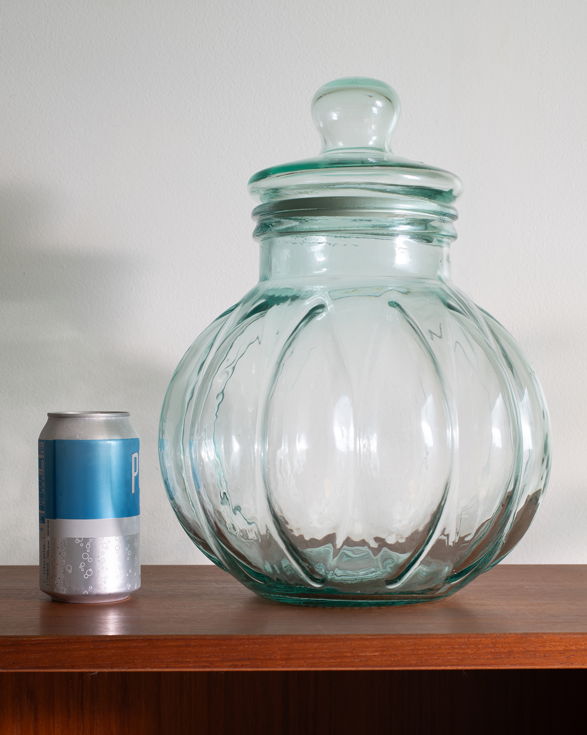 A large clear glass jar with a matching lid, featuring a ribbed, pumpkin-like design, displayed on a wooden shelf next to a soda can for scale.
