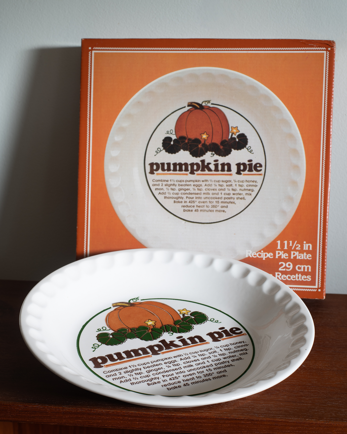A vintage-style white ceramic pumpkin pie plate with a printed orange, brown, and green pie graphic and text, shown in front of its orange retail box on a wooden surface against a gray wall.